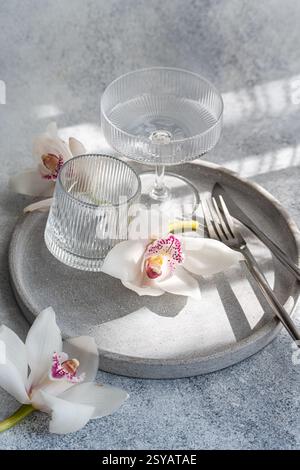 Une salle à manger raffinée avec verrerie côtelée, orchidées délicates et couverts élégants sur un plateau circulaire en béton, créant un cadre sophistiqué et serein Banque D'Images