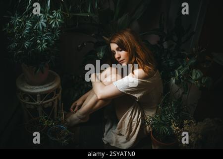 Une jeune femme s'assoit paisiblement parmi les plantes d'intérieur luxuriantes, entourée de verdure. Son expression sereine et sa posture détendue évoquent un sentiment de tranquillité Banque D'Images