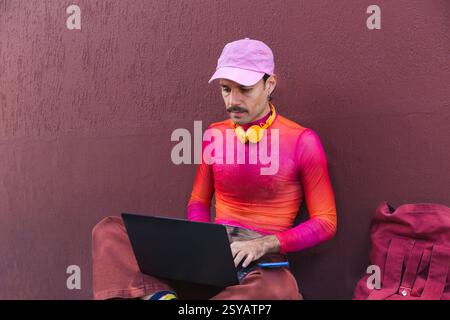 À Quito, en Équateur, un jeune Espagnol vêtu de rose travaille sur son ordinateur portable à l'extérieur contre un mur violet. Sa moustache et sa tenue décontractée et colorée e Banque D'Images