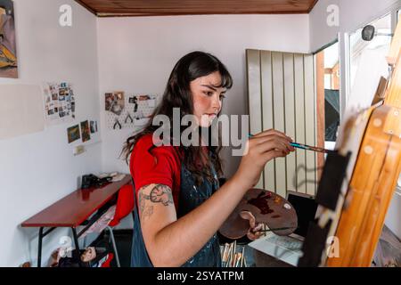 Une jeune femme artiste se concentre sur la peinture d'une toile dans son home studio. L'espace confortable est rempli de matériaux d'art et de lumière naturelle, créant un in Banque D'Images