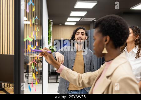 Une équipe discute des idées créatives tout en utilisant des notes adhésives colorées sur une planche de verre dans un bureau moderne. Ils s'engagent dans un brainstorming collaboratif pour f Banque D'Images