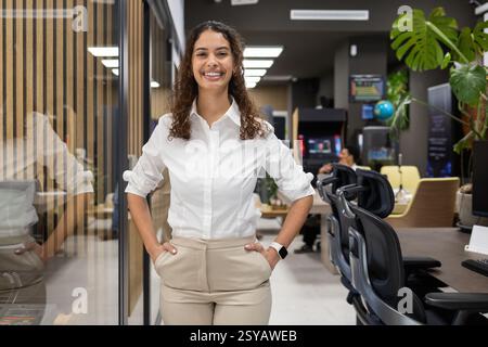Une femme dans une blouse blanche se tient confiante dans un bureau moderne. Son sourire reflète un comportement positif et professionnel, avec un décor et des œuvres élégantes Banque D'Images