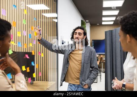 Les professionnels participent à une séance de brainstorming dans un espace de bureau moderne. Un homme fait des gestes vers des notes collantes colorées sur verre, favorisant la créativité Banque D'Images