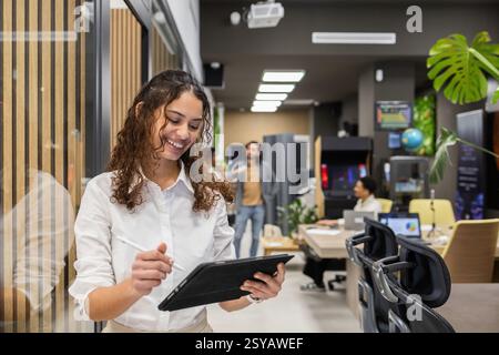 Un professionnel joyeux dans un bureau moderne occupé examine les informations sur une tablette. Les collègues travaillent en arrière-plan, créant une collaboration active et Banque D'Images