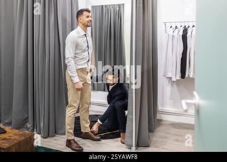 Un tailleur professionnel mesure un homme pour l'ajustement de pantalon dans une cabine d'essayage élégante. Le client se tient souriant devant un miroir, entouré d'une chemise Banque D'Images