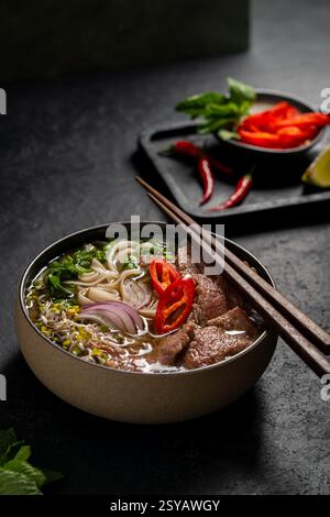 Pho de bœuf vietnamien traditionnel servi dans un bol, avec des tranches de bœuf, des nouilles, des oignons rouges, des piments, et un riche, bouillon clair, accompagné d'un si Banque D'Images