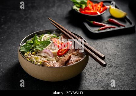 Soupe traditionnelle vietnamienne de nouilles Pho au bœuf dans un bol en céramique, garnie de tranches de bœuf, d'herbes, de piment rouge et de citron vert sur fond de pierre sombre. Banque D'Images