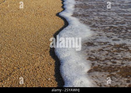 Gros plan de vagues douces et de mousse rencontrant une plage de sable sous la lumière dorée du soleil, capturant l'essence sereine et apaisante d'un paysage côtier. Banque D'Images