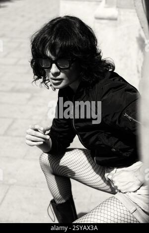 Une femme élégante aux cheveux désordonnés et aux lunettes de soleil respire la confiance dans ce portrait noir et blanc à contraste élevé. Vêtu de collants en filet et d'une bombe Banque D'Images