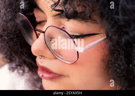 Gros plan détaillé d'une femme latine portant des lunettes. L'accent est mis sur leurs piercings, maquillage et cheveux bouclés, capturant un style unique et Banque D'Images