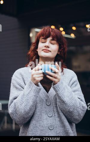 Une jeune femme aux yeux fermés savoure un moment avec une tasse de café. Elle porte un pull gris confortable, debout à l'extérieur sur la terrasse du café, créant une sere Banque D'Images