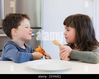 Deux enfants partagent joyeusement un repas à une table de cuisine, tenant des tranches de pizza. La scène capture leur bonheur et leur compagnie dans un kitch lumineux Banque D'Images