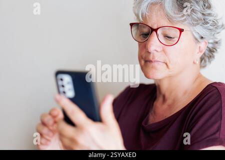 Femme senior avec les cheveux gris et les lunettes rouges à l'aide d'un smartphone. Elle est concentrée et engagée, assise confortablement dans un cadre intérieur bien éclairé avec neut Banque D'Images