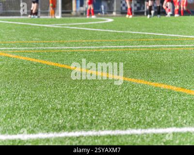 Gros plan d'un terrain de football à texture artificielle verte avec des lignes blanches et jaunes brillantes. Les joueurs flous dans des uniformes colorés se tiennent en arrière-plan n Banque D'Images