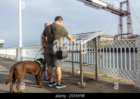 Un aveugle avec un chien-guide a lu un panneau d'information sur un pont surplombant une rivière pittoresque et des bâtiments lointains. La conception industrielle du pont ajoute Banque D'Images