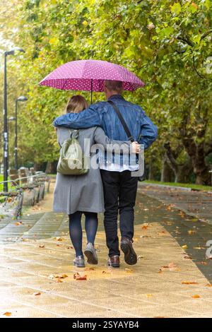 Un couple ayant une déficience intellectuelle se promène dans un parc sous un parapluie rose. La scène capture le romantisme et la tranquillité d'un jour de pluie wa Banque D'Images