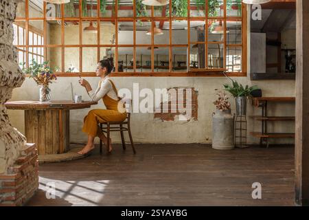 Une femme se livre au télétravail à l'aide d'un ordinateur portable sur une table circulaire en bois dans un café rustique et magnifiquement vieilli avec des murs de briques visibles et de grandes fenêtres Banque D'Images