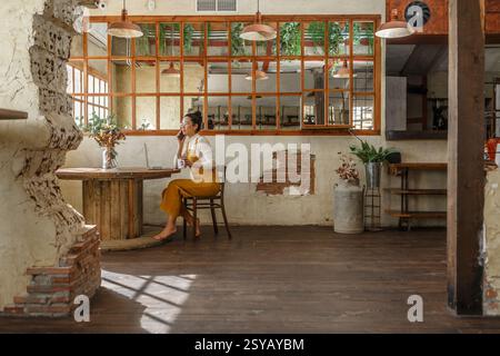 Une femme télétravaille dans un café de style vintage, en utilisant un ordinateur portable et un smartphone pour engager une conversation téléphonique. Banque D'Images