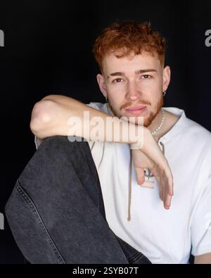 Un jeune homme aux cheveux roux et à la barbe, portant un sweat à capuche et un Jean, assis sur un fond noir. Son expression est réfléchie, ajoutant un artisti Banque D'Images