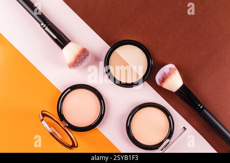 Pose plate de pinceaux de maquillage et de poudres pour le visage sur un fond tricolore vibrant. Les surfaces contrastantes orange, blanches et brunes soulignent la beauté p Banque D'Images