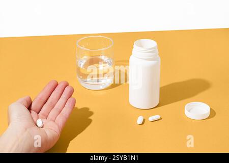 Une main tient une pilule blanche au-dessus d'une surface jaune, à côté d'une bouteille ouverte, des pilules dispersées et un verre d'eau. La scène suggère la santé quotidienne ou moi Banque D'Images