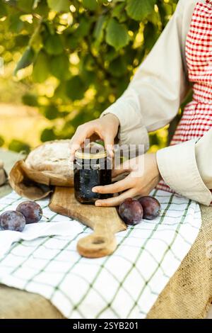 Mains préparant des canapés à l'extérieur sur une planche de bois. Les prunes fraîches, la confiture maison et le pain artisanal sur une nappe rustique créent un SC confortable et délicieux Banque D'Images