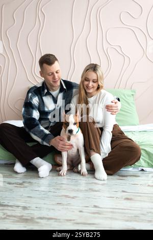 Un jeune homme et une jeune femme sont assis sur le sol à la maison, souriant joyeusement alors qu'ils interagissent avec leur chien blanc et bronzé. Ils affichent un corps affectueux Banque D'Images