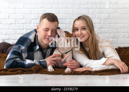Un jeune couple partage un moment d'amour avec leur chien blanc et brun énergique, allongé sur une couverture confortable à la maison, souriant. Banque D'Images