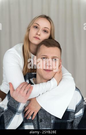 Un jeune homme est assis comme une femme l'embrasse par derrière, à la fois calme et affectueux dans leur cadre confortable de maison. Banque D'Images