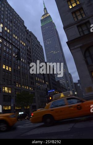 Le crépuscule tombe sur Manhattan alors que l'emblématique Empire State Building s'illumine, vu entre les grands bâtiments de la ville avec un taxi jaune qui passe. Banque D'Images