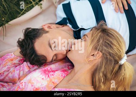 Un couple aimant profite d'un moment romantique à l'extérieur, allongé sur une couverture dans l'herbe. Leur baiser tendre et leur posture détendue reflètent l'amour, l'intimité, et Banque D'Images