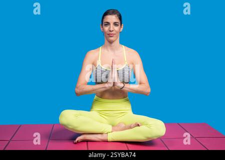 Professeur de yoga méditant dans la pose de lotus sur un tapis rose, montrant les mains en position de prière sur un fond bleu Banque D'Images
