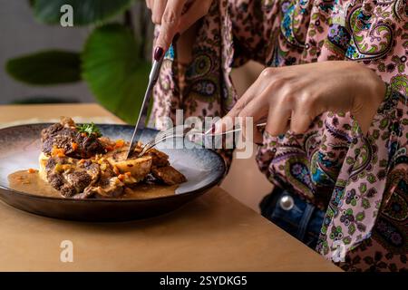 Gros plan d'une femme coupant dans une assiette gourmande d'ossobuco avec de la moelle osseuse, servie dans une sauce riche avec des légumes. Une expérience gastronomique raffinée showca Banque D'Images