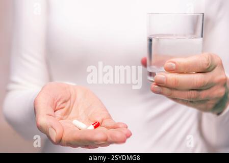 La personne se prépare à prendre des médicaments en tenant des capsules dans une main et un verre d'eau dans l'autre tout en restant debout à l'intérieur. Banque D'Images