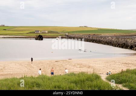 Les gens à la plage par une journée ensoleillée à côté de Churchill Barrier 3 entre Glimps Holm et Burray, Orcades. Bloquer le navire dans l'est du détroit de Weddell en arrière-plan. Banque D'Images