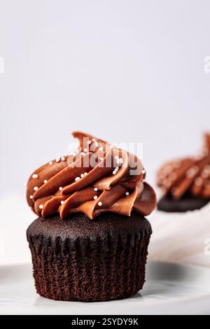 Gros plan d'un cupcake au chocolat noir recouvert d'un glaçage au chocolat tourbillonnant et de délicates perles blanches sur fond blanc minimaliste. Banque D'Images