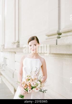 Une mariée pose élégamment à l'extérieur d'une grande structure, tenant un bouquet luxuriant de fleurs dans la douce lumière de l'après-midi. Banque D'Images