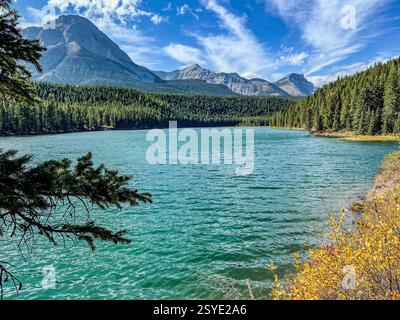 Un lac turquoise se trouve au pied d'une chaîne de montagnes imposante, entouré de forêts denses à feuilles persistantes. Le ciel est bleu vif avec des nuages tordus s'étirent Banque D'Images