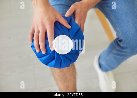 Gros plan de la main de l'homme tenant le sac de glace appliquer la compresse froide à la jambe assise sur la chaise à l'intérieur Banque D'Images
