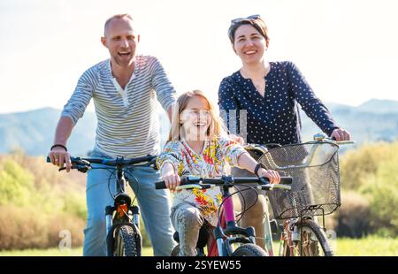 Sourire père et mère avec fille pendant l'été à vélo en plein air. Ils apprécient la convivialité sur la prairie verte de hautes herbes. Bonne parentalité et Banque D'Images