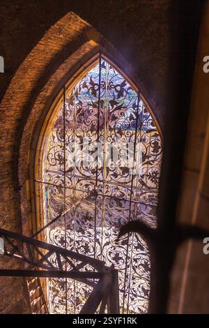 Une photo en bas angle d'un vitrail orné avec des motifs complexes et une ossature métallique, encadrée par une arche en pierre et une partie d'escalier. Banque D'Images