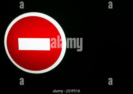 Un panneau circulaire rouge vif avec une barre horizontale blanche est éclairé sur fond sombre, signalant l'interdiction d'accès aux véhicules dans une zone réglementée d Banque D'Images