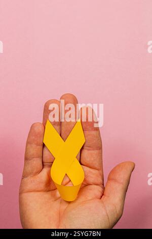 Une main féminine tient un ruban de sensibilisation jaune sur un fond rose. Cela symbolise la solidarité et l’espoir dans la lutte contre le cancer et son chall Banque D'Images