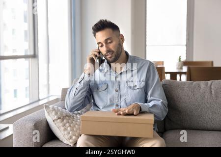 Homme assis sur le canapé tenant paquet parlant sur smartphone Banque D'Images