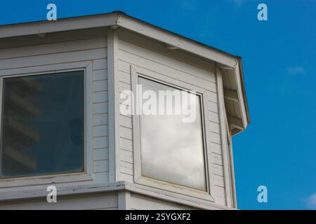 Baies vitrées. Façade d'une maison rurale en bois blanc avec deux fenêtres dans la journée. Coin d'un vieux bâtiment abandonné contre ciel bleu. Toit plat. Banque D'Images
