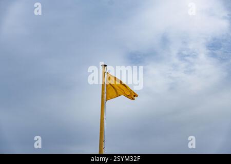 Un drapeau jaune flotte dans le ciel. Le ciel est principalement bleu avec quelques nuages. Le drapeau est légèrement déchiqueté et usé Banque D'Images