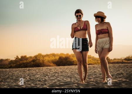 Deux jeunes femmes en bikinis et tenues d'été décontractées marchant pieds nus sur la plage au coucher du soleil, souriantes et profitant de l'atmosphère chaleureuse du soir. Banque D'Images