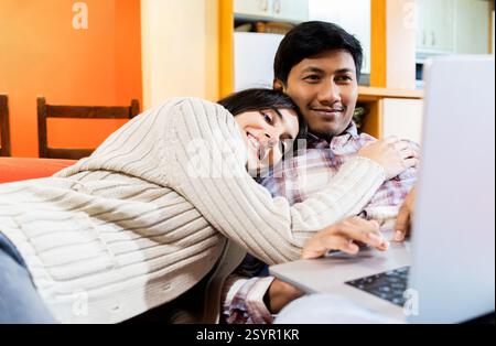 Heureux couple profite d'un moment confortable ensemble sur le canapé, avec la femme embrassant son partenaire pendant qu'il utilise un ordinateur portable. ils partagent un atmo chaleureux et détendu Banque D'Images