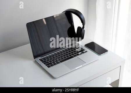 Ordinateur portable argenté ouvert, casque sans fil noir et smartphone sur un bureau blanc Banque D'Images