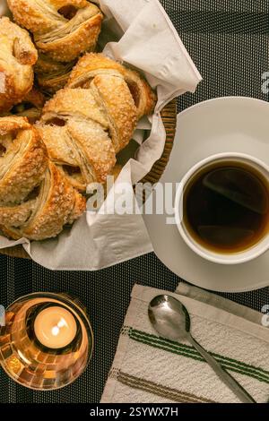 Une scène de petit déjeuner confortable avec strudels de pommes, pâtisseries dans un panier, une tasse de thé et une bougie allumée sur un napperon tissé Banque D'Images